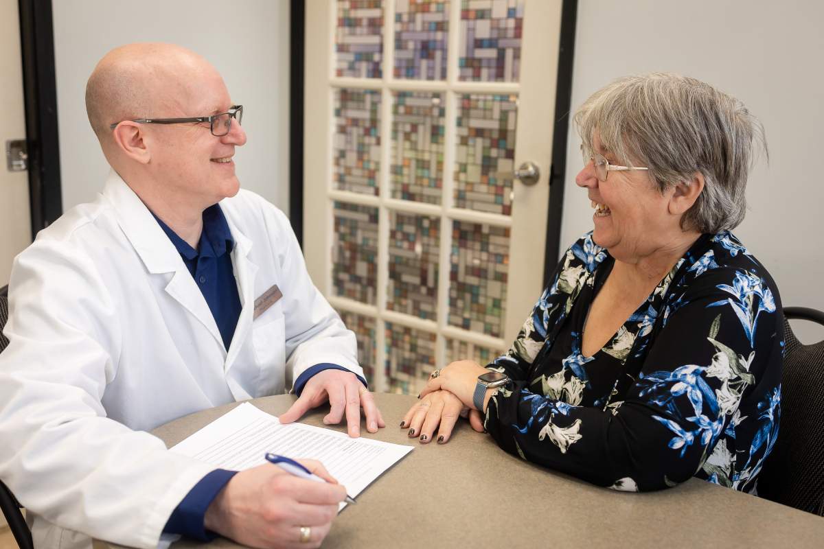A pharmacist conversing with an elderly woman during a consultation. They are seated at a table, with the pharmacist holding a pen and taking notes.